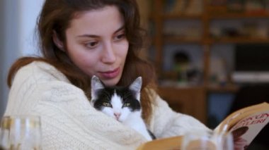 Tatil sezonunun ruhunun ortasında, kablo örgü süveter giyen dalgalı esmer saçlı genç bir kadın, Noel temalı bir kitaptan teselli buluyor, siyah-beyaz kedi arkadaşı