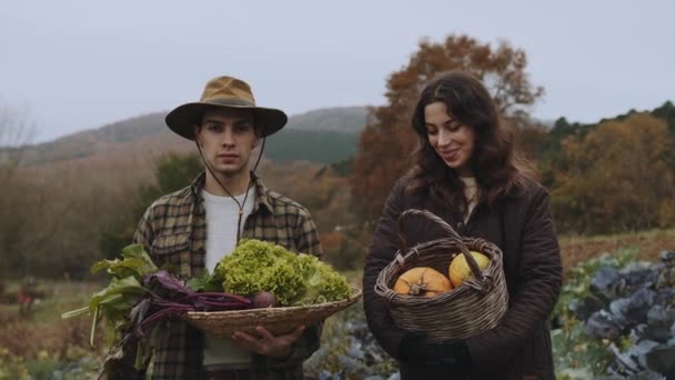 収穫時間 カップルは誇らしげに新鮮な農場の農産物を表示します 男と女が野原に立って 新鮮な野菜のバスケットを持っている 帽子と平らなシャツの男は緑を保持する一方 女性は — ストック動画