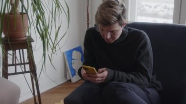 Evde Dijital Nişan: Smartphone Kullanımında Özümsenmiş Özel Bir Kanepe Üzerinde. Bir insan kendini akıllı telefonuna kaptırır, karanlık bir kanepede rahat bir şekilde oturur, etrafında rahat bir ev içi vardır.