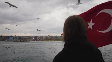 Sahne bir feribotta çekiliyor ve bir şahıs elinde belirgin bir hilal ve yıldızla büyük bir ulusal bayrak tutuyor. Rüzgarda dalgalanan bayrak, gri bulutlarla dolu bir arka planda