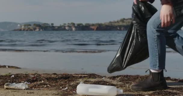 展望中 一位身穿深色夹克和牛仔裤的年轻女子蹲在海滩上 将垃圾收集到一个黑色的垃圾袋中 她的脸看不见 把观众的注意力集中在服务的动作上 — 图库视频影像
