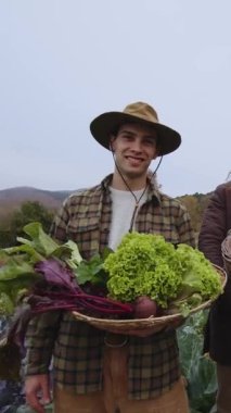 Neşeli genç bir adam, yaklaşık yirmili yaşlarda, elinde taze çiftlik ürünleriyle dolu bir sepet tutarak yeşil bir tarlada duruyor. Sepette canlı yeşil marul ve bol renkli salata var.