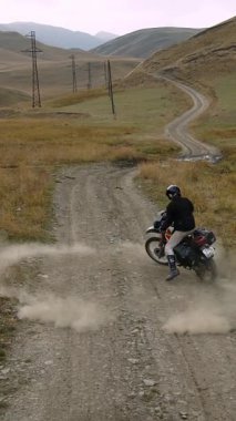 Cesur bir manevranın ortasında yakalanan siyah başlıklı motosikletli, çakıllı bir dağ yolunda dar bir çember dönüşü gerçekleştirir. Bisiklet, bir açıyla, kalın bir bulut fırlatır.