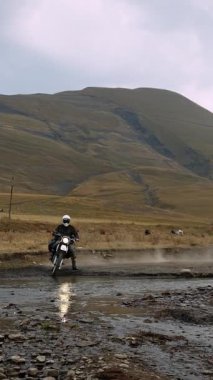 Dağlık arazinin göbeğinde korkusuz bir motosikletçi, nehirden karşıya geçerken motoru dinamik sıçramalarla suyu kesiyor. Beyaz bir miğfer ve koruyucu giysiler giymişti.