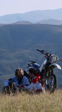Bu sakin sahne, sonbahardan fısıldayan kuru otlarla dolu bir arazide oturan iki motosikletlinin yolculuklarına ara verişini resmediyor. Off-road motosikletleri onların yanında duruyor.