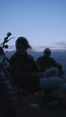 Alacakaranlık dağ kampını kucaklarken, kamp ateşinin ince ışığıyla aydınlanan bir grup arkadaş, dingin alacakaranlığın tadını çıkarıyor. Motosikletlerin siluetleri maceralı bir güne dönüştüğünü gösteriyor.
