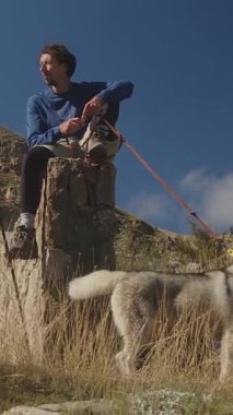 Dikey bir sinemada, mavi giyinmiş genç bir erkek tırmanıcı, bir tırmanma halatı emniyete alarak engebeli bir taş sütunun üzerinde oturur, tabanında tüylü, beyaz bir köpek,