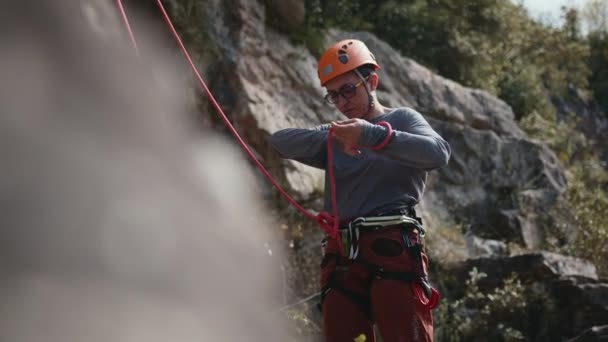 一个戴着头盔和带着安全带的专注的登山者正在精心地准备他的攀爬绳 他的注意力集中在手头的任务上 他站在一块朦胧的岩石上 攀爬着 — 图库视频影像