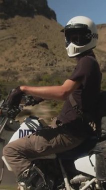Beyaz kasklı, siyah gömlekli ve eldivenli bir motosikletçi kurak bir dağ manzarasında motosiklet sürüyor. Arkaplanda engebeli, çorak tepeler ve berrak yeşil yapraklar yer alıyor.