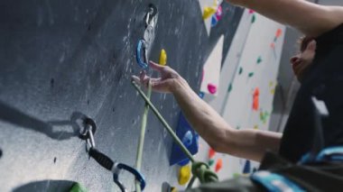 Kapalı alan tırmanma duvarına tutunan bir dağcının yakın plan çekimi. Görüntü, tırmanışçıların güvenlik halatı ile korunan duvarı aşma tekniği ve hassasiyetine odaklanıyor. Şey...