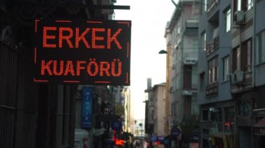 İstanbul 'un hareketli caddesinde berber dükkanı için parlak bir tabela var. Tabelada Mens Kuaför yazıyor ve makas görüntüsüyle yer değiştiriyor. Cadde çok katlı binalarla dolu.