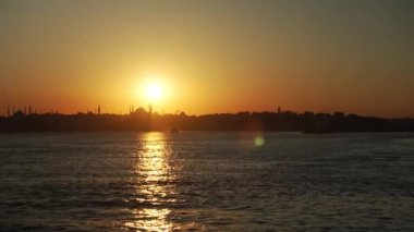 Gün batımında sakin bir an, insan siluetleriyle dolu bir feribot. Feribot ön planda, altın güneş arka planda batarken, sıcak ve huzurlu bir şekilde