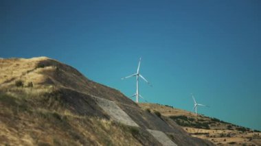 Güneşli bir günde açık mavi gökyüzüne karşı kayan tepelerdeki rüzgar türbinlerinin panoramik görüntüsü. Ön planda seyrek bitki örtüsüne sahip kayalık bir yamaç yer alırken, arka planda bir dizi