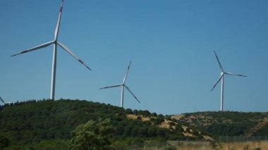 Sakin bir manzarada, berrak mavi bir gökyüzünün altındaki bir yamaçta yavaşça dönen büyük rüzgar türbinleri bulunur. Bu etkileyici yapılar yenilenebilir enerji projesinin bir parçasıdır.