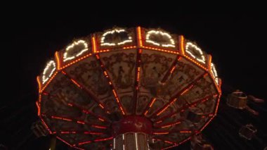 Aydınlatılmış bir salıncaklı atlıkarıncayı karanlık bir gökyüzüne doğru hareket ederken dinamik bir çekim. Atlıkarınca, şenlikli ve büyülü bir atmosfer yaratan karmaşık tasarımlar ve canlı ışıklar içeriyor. Parlak