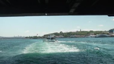 Tarihi bir köprünün altından geçen bir tekne berrak mavi sularda görülebiliyor. Kentsel ufuk çizgisi arka planda görülebilir, modern şehirle şehir arasındaki zıtlığı vurgular.