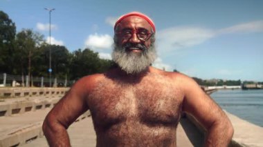 Kas yapısı ve bronzlaşmış derisi olan atletik yaşlı bir Türk, açık havada, bir rıhtımın yanında duruyor. Kırmızı bir saç bandı ve gözlük takıyor. Gülümsüyor ve mutluluk saçıyor. Göğsü.