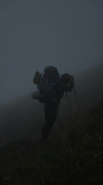 Bir yürüyüşçü, dik, çimenli bir yamaca tırmanırken, kalın bir sise karşı siluet şeklindedir. Karanlık, sisli koşullar atmosferik ve karamsar bir sahne yaratıyor.