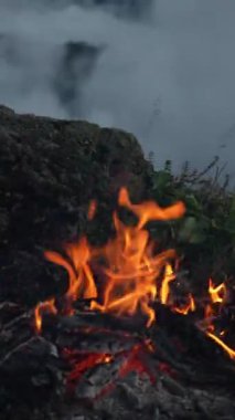 Engebeli bir dağ yamacında parlayan bir kamp ateşinin yakın görüntüsü. Alevler, kayan sis ve uzak bulutların zemininde dans ediyor. Ateşin yoğun parlaklığıyla çelişiyor
