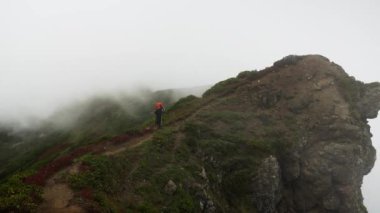 Yalnız bir yürüyüşçü, yoğun sis altında dar bir dağ sırtı patikası boyunca yürür. Yürüyüşçü geniş turuncu bir sırt çantası ve engebeli arazide dikkatlice gezinen yürüyüş direkleriyle donatılmıştır. Şey...