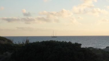 Güçlü okyanus dalgaları, batan güneşin aydınlattığı dağınık bulutlarla dolu pastel renkli bir akşam gökyüzünün altında yüzeye çarpar. Ufukta küçük bir balıkçı teknesi görünüyor.