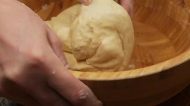 Ahşap bir kasede hamur yoğuran güçlü erkek ellerinin detaylı bir yakın çekimi. Bir bileğinde siyah yıldız dövmesi var ve parmakları un tozuyla kaplanmış. Eller yumuşak hamura bastırır