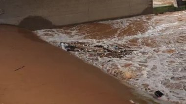 Kahverengi fırtına suyu, köpük, plastik atık ve ağaç dallarıyla dolu bir köprünün altından akıyor. Görülebilir kirlilik ve kentsel akış, sağanak yağmurun ardından yetersiz drenaj ve çevresel ihmalkarlığı yansıtıyor