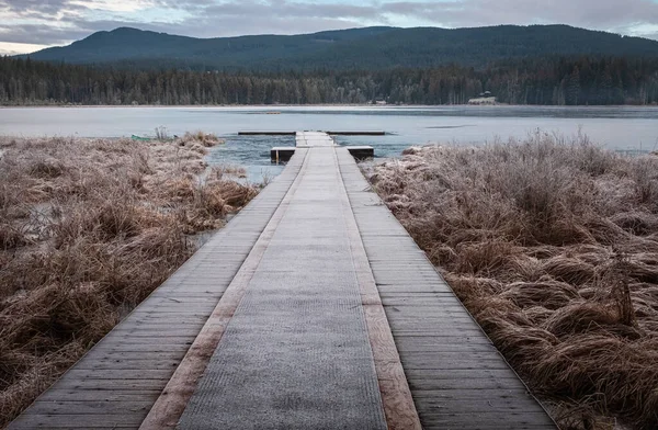 Wooden pier on lake site facing a beautiful mountain on a misty winter morning. Cold winters morning on a forest lake. Frosty calm landscape. Natural tranquil atmosphere for relaxation
