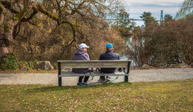 Yaşlı bir çift parkta bir bankta dinleniyor. Yaşlı bir çiftin gölün kıyısındaki ahşap bir odada oturduğunu gördüm. Vancouver Canada 'da 30 Mart 2023' te bir bankta dinlenen yaşlı bir çift. Sokak görüntüsü, seçici odak
