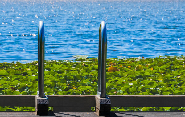 Ladder on deck above rippled blue water in bright sunlight. Lake background with dock ladder. A Stainless Steel Ladder Leads to the Water at outdoor summer park. Stainless steel grab bars for ladders