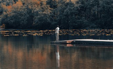 Young man standing alone on edge of footbridge and relaxing at lake. Man standing alone on the end of a jetty, looking over a forest lake. Man spending time with nature. Copy space, travel photo. clipart