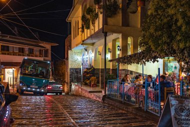 Kafe masaları ve yemek yiyen turistlerle dolu eski bir cadde. Arnavutluk 'un Cirokaster kentindeki sokak restoranları. Arnavutluk 'un eski kenti 27 Ekim 2023' te gece çalışan açık hava restoranları