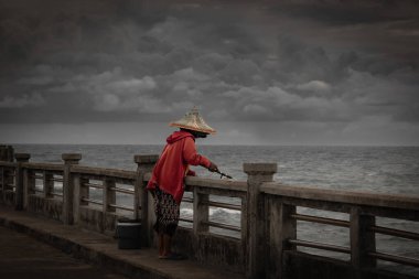 Tayland 'da oltayla balık avlayan kadınlar. Fırtına sırasında limanda balık tutan kırmızı ceketli Asyalı kadın. Bulutlu bir günde yağmur mevsiminde balık tutmak. Palmiye yaprağı şapkalı kadın.