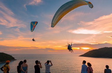 Phuket Tayland 'da mavi gökyüzü ile paraşütle atlamak. Paragliderler gökyüzünde uçuyor. İnsanlar gün batımında paraşütçülerin uçuşunu izliyor. Tayland yamaç paraşütü okulu. 8 Temmuz 2024