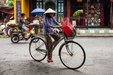 Hoi An Vietnam 'da bisiklet süren koni şapkalı yaşlı bir kadın. Geleneksel bambu şapkalı Vietnamlı kadın Hoi An Antik Kasabası boyunca bisiklet sürüyor. Seyahat fotoğrafı, sokak fotoğrafı - 21 Mayıs 2024