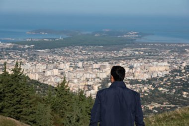 Young man standing on top of cliff in mountain and enjoying view of Vlore city in Albania. A man looking down on the city at the valley,standing on the top of the hill. Travel photo clipart