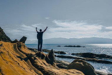 Sırt çantalı bir adam güneşli bir günde uçurumun kenarında kollarını kaldırıyor. Dışarıda huzurlu bir anın tadını çıkarıyorum. Sağlık, sağlık, seyahat ve zihinsel sağlık kavramı.