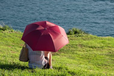Nehir ya da deniz kenarındaki kırmızı şemsiye altında yeşil yaz çayırında dinlenen bir kadın. Güneşli bir günde parkta şemsiye altında oturan genç kadın. Doğal bir hafta sonu.
