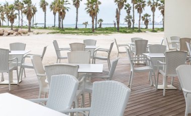 Deniz kenarında bir yaz günü deniz kenarında beyaz masalar ve sandalyeler yerleştirildi. Palmiye ağaçlarıyla dolu kumlu sahil kenarındaki restoran kafesinin boş terası. Yaz tatili konsepti. Seyahat fotoğrafı.