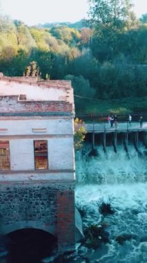 Nehir kenarında terk edilmiş eski bir değirmen. Nehrin kıyısında barajı olan harap bir değirmen. Dikey video.