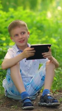 Bir çocuk parkta yürüyor ve bir tablette kulaklıkla müzik dinliyor. Dikey video.