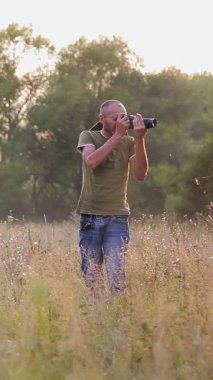 Erkek fotoğrafçı vahşi doğada gün batımında güzel bir manzara yaratıyor. Açık havada çalışan bir fotoğrafçı. Fotoğraf avı. Akşam güneşinde işini yapan bir fotoğrafçı. Dikey video.