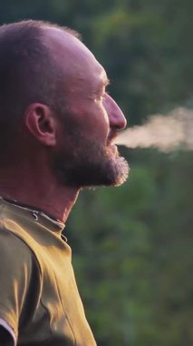 Vahşi doğada sigara içen biri. Doğada gün batımında sigara içen adam. Gezgin sigara içiyor ve doğada dinleniyor. Dikey video.