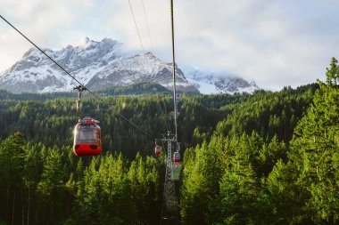 Kırmızı teleferik yemyeşil çamlarla çevrili, kalın bulutlar, kar kaplı dağların arkasında, Kriens, İsviçre.
