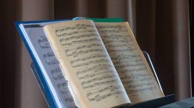 Piyanonun üzerinde notalı açık bir klasör var. Perdelerin arka planında eski piyano notaları