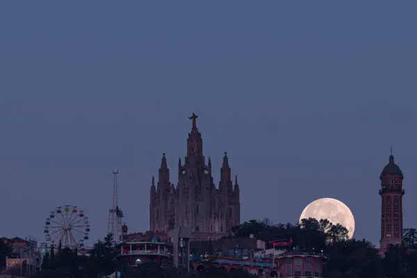 Tibidabo, Barcelona 'daki İsa' nın Kutsal Kalbi tapınağı ile antik bir su tankı olan su kulesi arasındaki dolunay batımı. Tapınağın solunda dağdan sonra adlandırılan eğlence parkını görebilirsiniz, Tibidabo 'nun bulunduğu yer.