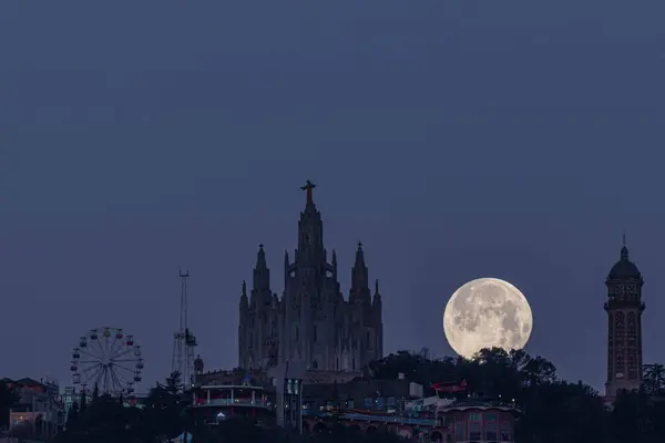 Tibidabo, Barcelona 'daki İsa' nın Kutsal Kalbi tapınağı ile antik bir su tankı olan su kulesi arasındaki dolunay batımı. Tapınağın solunda dağdan sonra adlandırılan eğlence parkını görebilirsiniz, Tibidabo 'nun bulunduğu yer.