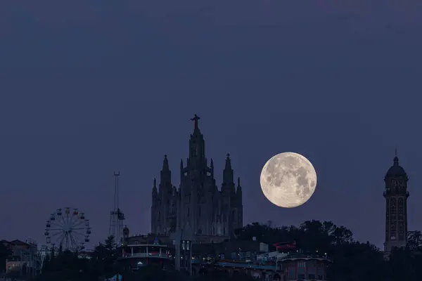 Tibidabo, Barcelona 'daki İsa' nın Kutsal Kalbi tapınağı ile antik bir su tankı olan su kulesi arasındaki dolunay batımı. Tapınağın solunda dağdan sonra adlandırılan eğlence parkını görebilirsiniz, Tibidabo 'nun bulunduğu yer.