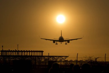 Bir uçağın inişi silueti. Gökyüzü tamamen turuncu çünkü güneş batıyor ve şimdiden çok alçak. Uçağın altında büyük siyah bir anten de görebilirsiniz..