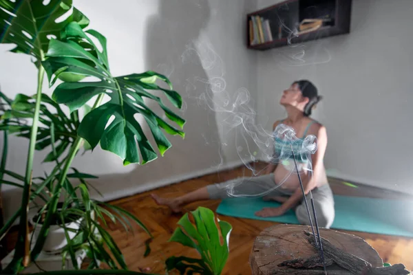 stock image Pregnant woman sits on a yoga mat in a meditation pose. Pregnant woman doing yoga exercises at home. Incense room for relaxation during yoga